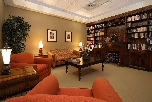 A cozy living room with two orange armchairs and a striped sofa arranged around a dark wooden coffee table. The room features a large wooden bookshelf with various books and decorative items, a fireplace, two table lamps on side tables, framed artwork on the wall, and a tall green plant in the corner.