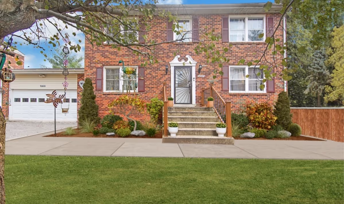Front exterior view of a two-story brick house with a white door, stairs leading up to the entrance, and several windows with shutters. There is a garage on the left side and landscaped bushes and plants around the house. A tree branch with hanging decorations is visible in the foreground.