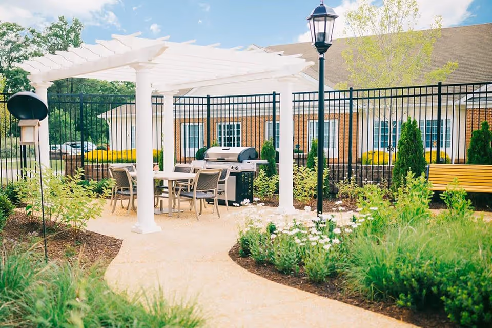 Outdoor patio area with a white pergola covering a table and chairs, a barbecue grill, a black metal fence, a lamppost, and landscaped garden beds with flowers and shrubs in front of a brick building.