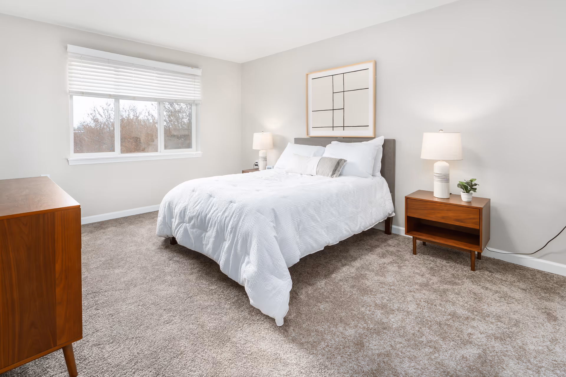A bright bedroom with a large window covered by white blinds, a bed with white bedding and pillows, two wooden nightstands each with a lamp, a small plant on one nightstand, a wooden dresser, and a framed abstract artwork on the wall above the bed.