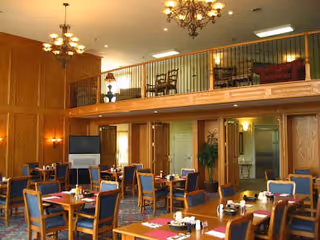 Dining room with multiple tables and chairs, wood-paneled walls, chandeliers, and an upper mezzanine seating area.