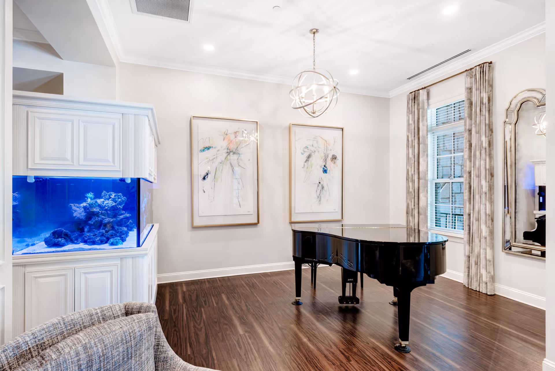 A bright room with a black grand piano on dark wooden flooring, two abstract paintings on the wall, a large window with patterned curtains, a tall mirror, and a built-in white cabinet with a blue-lit aquarium. A gray upholstered chair is partially visible in the foreground.