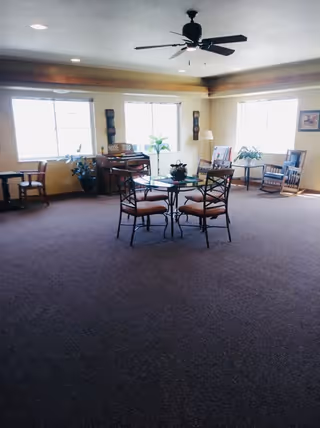 A spacious room with carpeted floor and large windows letting in natural light. The room features a ceiling fan, a round glass table with four chairs in the center, several potted plants, a piano against the far wall, and additional seating including wooden chairs and a small table near the windows.