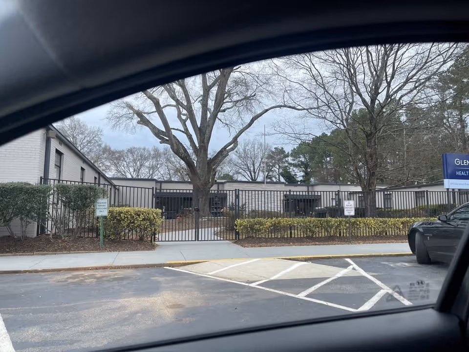 View from inside a car of the gated front of Glenwood Health Center, a low single-story building with large leafless trees.