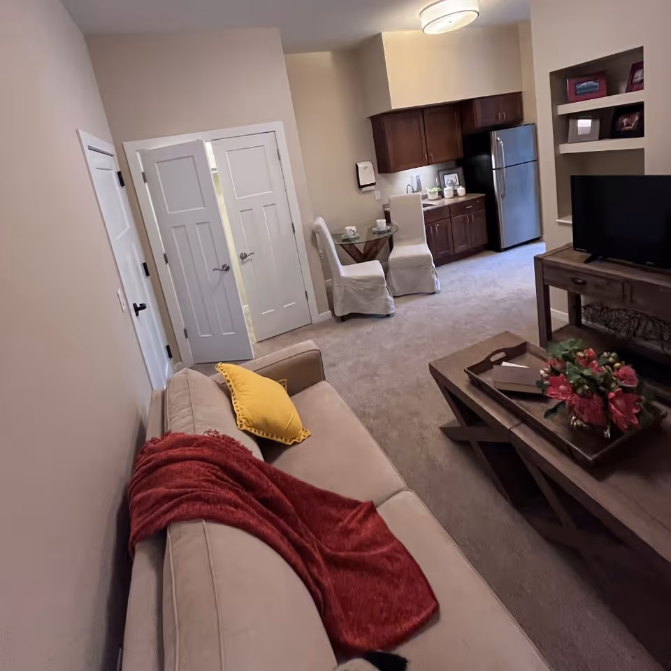 Interior view of a cozy living space in an assisted living facility featuring a beige sofa with a red throw blanket and yellow pillow, a wooden coffee table with a flower arrangement, a TV on a wooden stand, a small dining area with two white-covered chairs and a glass table, and a kitchenette with dark wood cabinets and a stainless steel refrigerator.