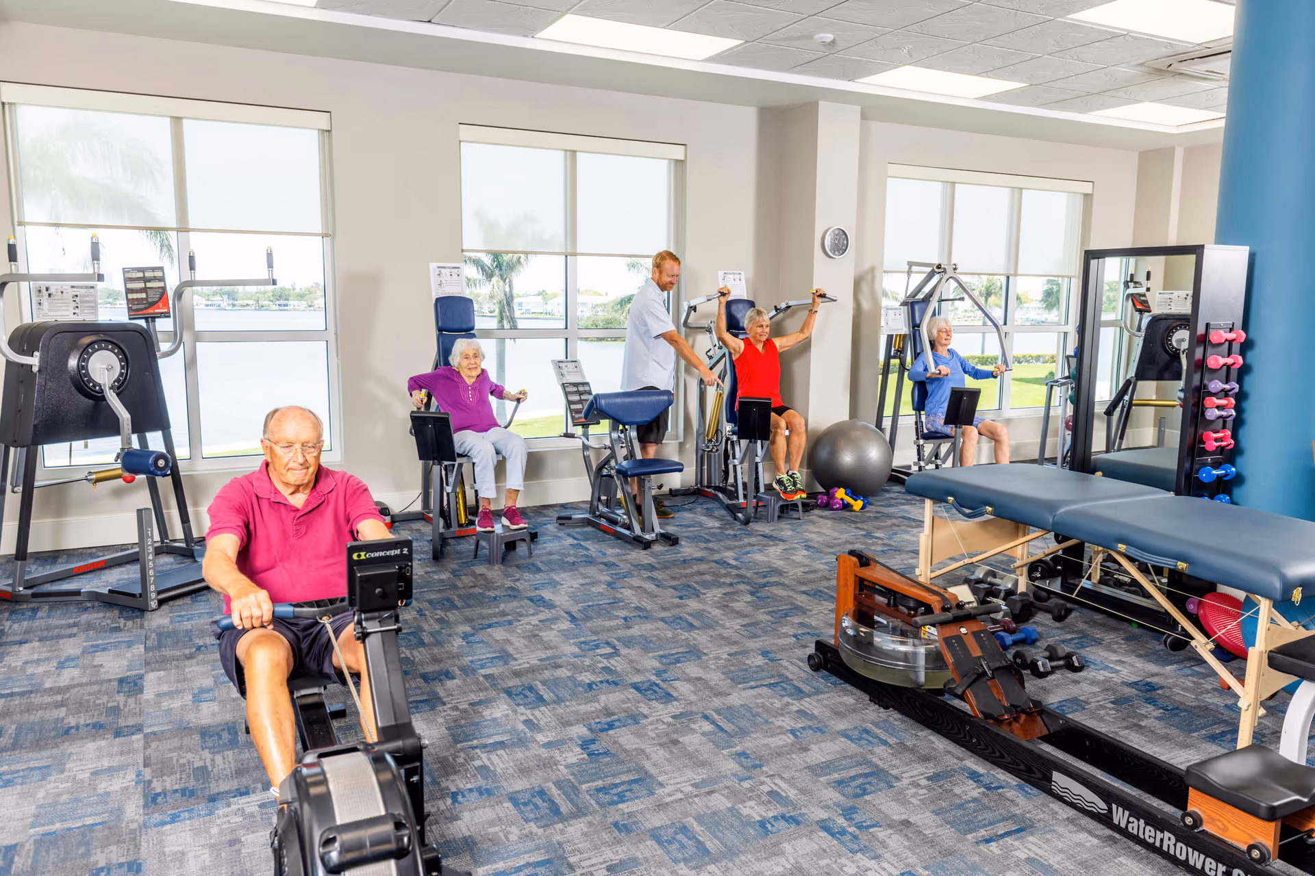 A bright fitness room with large windows showing an outdoor view. Four elderly individuals are using various exercise machines, assisted by a male staff member. The room has blue patterned carpet, exercise equipment including a rowing machine, weight machines, dumbbells, and a massage table.