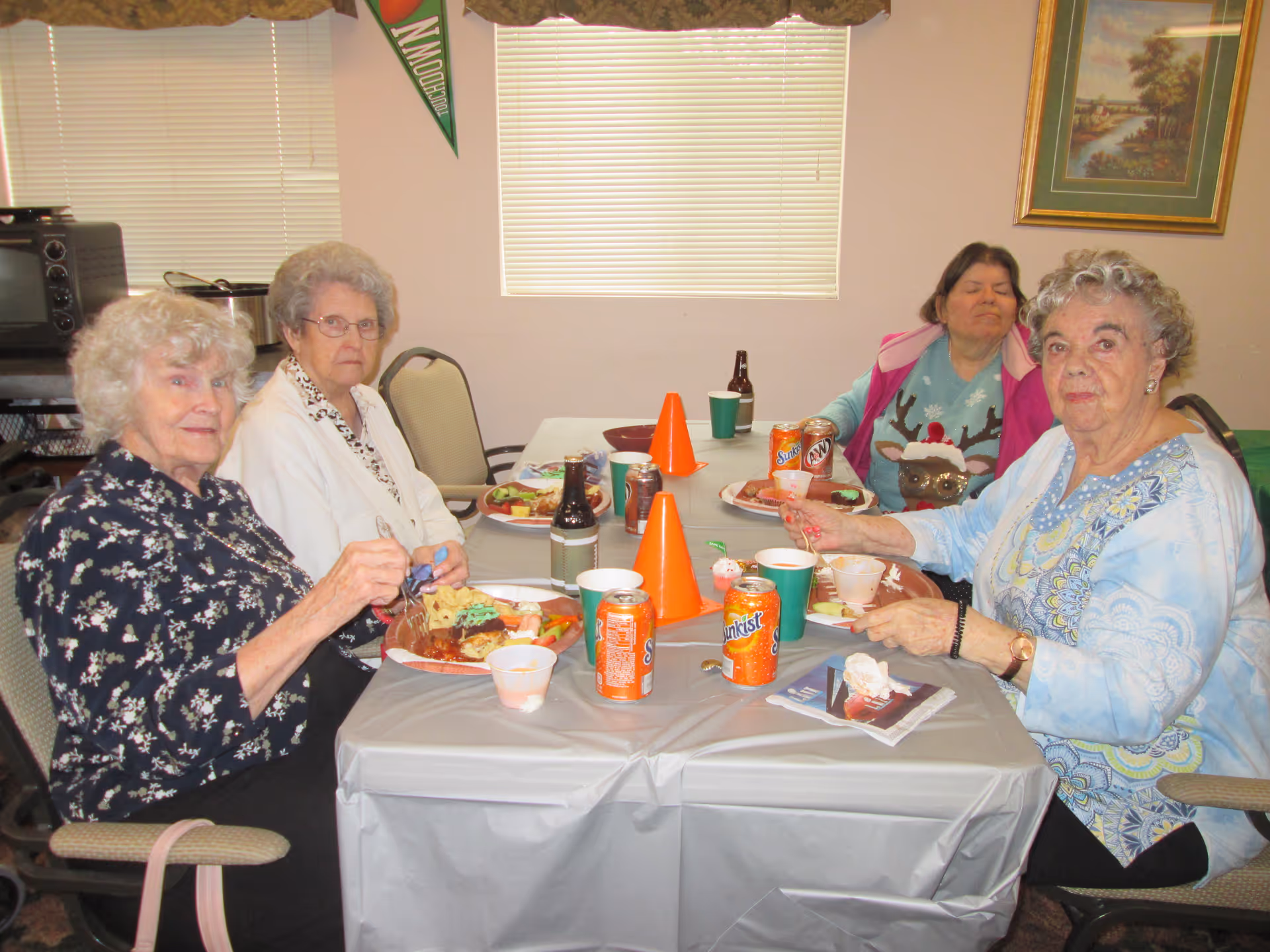 Four older adults sit around a table in a dining room eating a meal with plates, cups and soda cans.