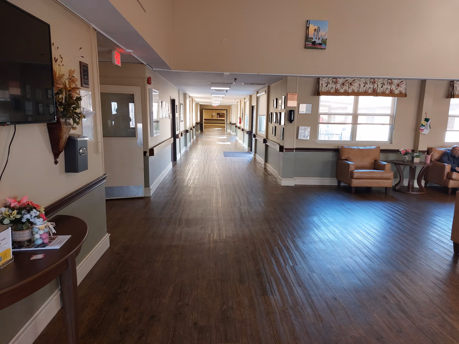 A long hallway in a nursing and rehabilitation facility with wooden flooring and beige walls. On the left side, there is a mounted TV above a small table with flowers and informational pamphlets. The hallway has several doors and handrails along the walls. On the right side, there is a seating area with brown leather chairs, a small table with flowers, and large windows with floral valances. A person is sitting in one of the chairs.