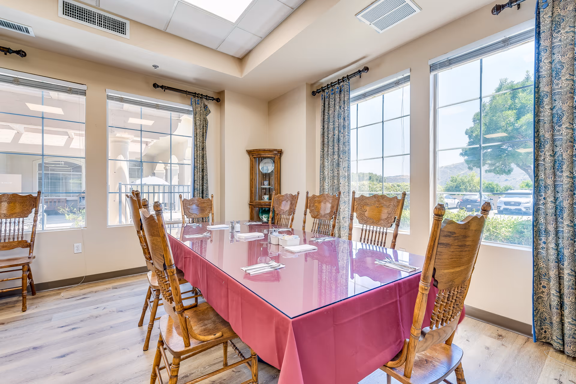 A bright dining room with large windows letting in natural light. There is a rectangular table covered with a red tablecloth and a glass top, surrounded by wooden chairs. The table is set with napkins, silverware, salt and pepper shakers, and a small condiment holder. Blue patterned curtains frame the windows, and a wooden corner cabinet with decorative plates is visible in the corner. Outside the windows, trees and parked cars can be seen.