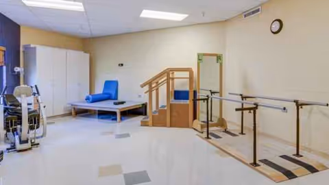 Indoor rehabilitation room with parallel bars, stairs, mats, and exercise equipment.