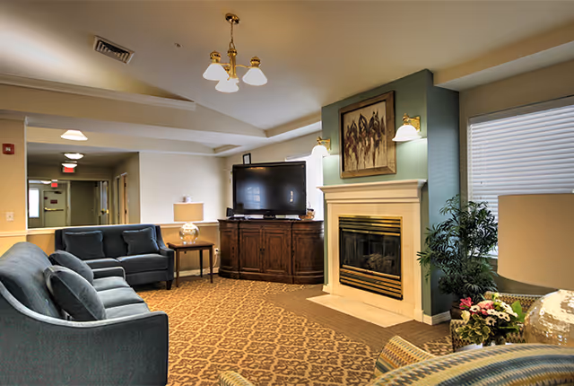 A cozy senior living facility common area with blue upholstered sofas, a wooden cabinet holding a flat-screen TV, a fireplace with a painting above it, a patterned carpet, and a potted plant near a window with blinds.
