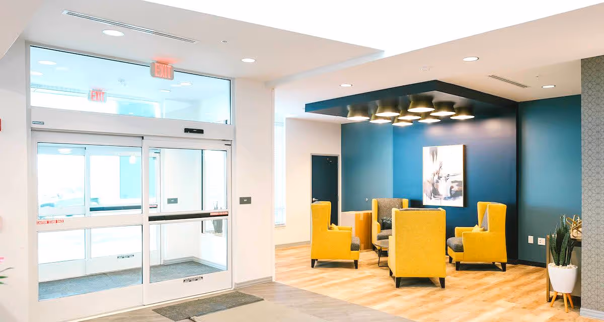 Bright and modern senior living facility lobby area with automatic glass entrance doors on the left and a seating area on the right featuring four yellow armchairs arranged around a small table, set against a dark teal accent wall with a piece of abstract artwork and overhead lighting fixtures.