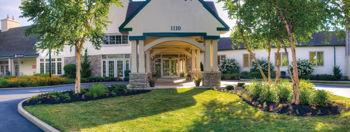 Front exterior view of a senior living facility named Lions Gate with a covered entrance, stone pillars, well-maintained landscaping including green grass, trees, and flower beds, and a building number 1110 displayed above the entrance.