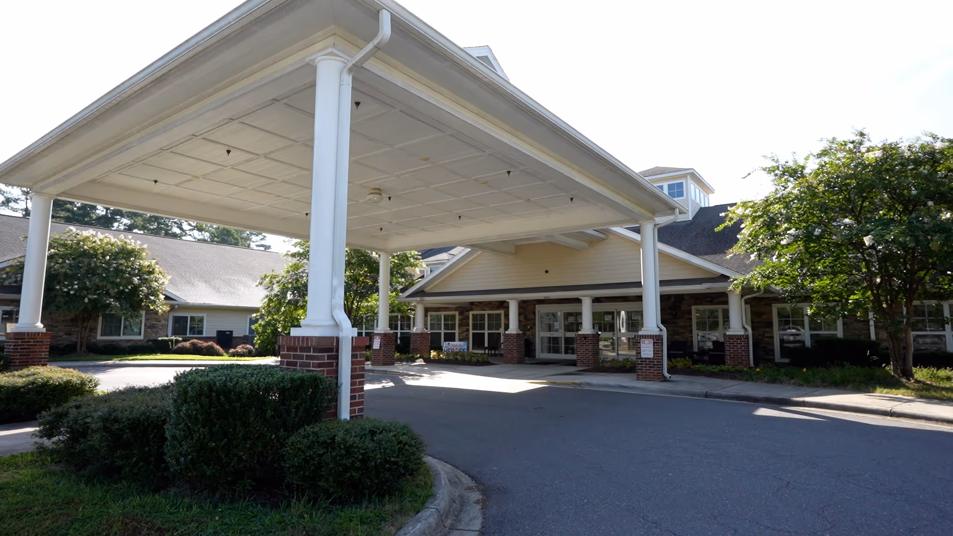 Entrance of The Pavilion Health Center at Brightmore featuring a covered drop-off area supported by white columns with brick bases, surrounded by greenery and trees under a clear sky.