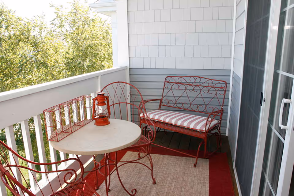 A small outdoor balcony with red metal furniture including a round table, two chairs, and a bench with a striped cushion. There is a red lantern and a red wire basket on the table. The balcony has white railing and overlooks green trees.