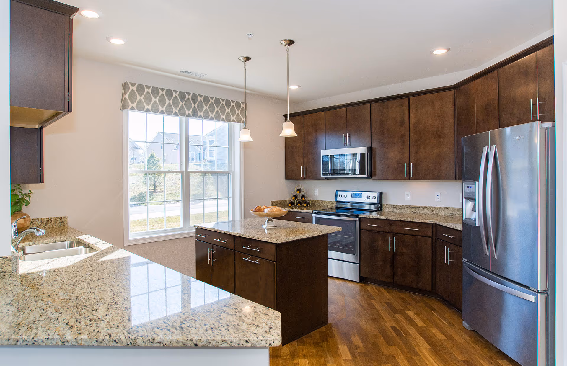Bright kitchen with dark wood cabinets, granite countertops, a central island, stainless steel appliances including a refrigerator, oven, and microwave, and a large window with a patterned valance letting in natural light.