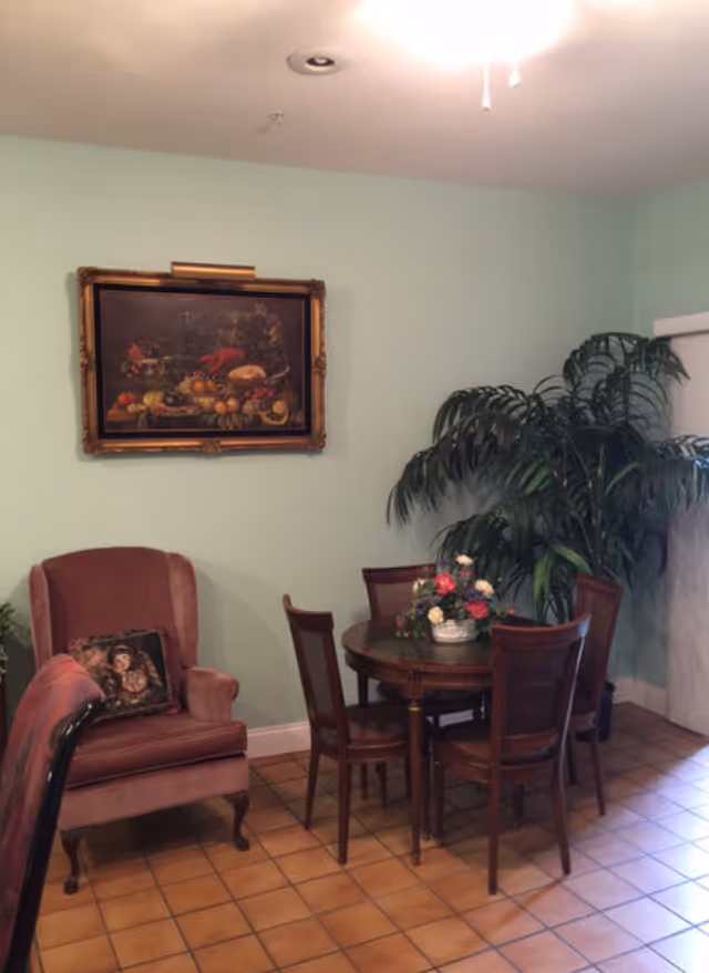 A cozy corner of a room with a round wooden table surrounded by four wooden chairs. A floral centerpiece is on the table. To the left, there is a plush armchair with a decorative pillow. A large potted plant is in the corner near a window with blinds. A framed painting of a still life with fruits hangs on the light green wall above the armchair. The floor is tiled with brown square tiles.