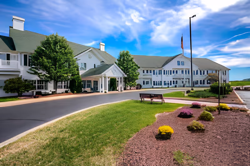 Front exterior of a large white senior living building with a covered entrance, landscaped lawn, benches, and a flagpole under a blue sky.