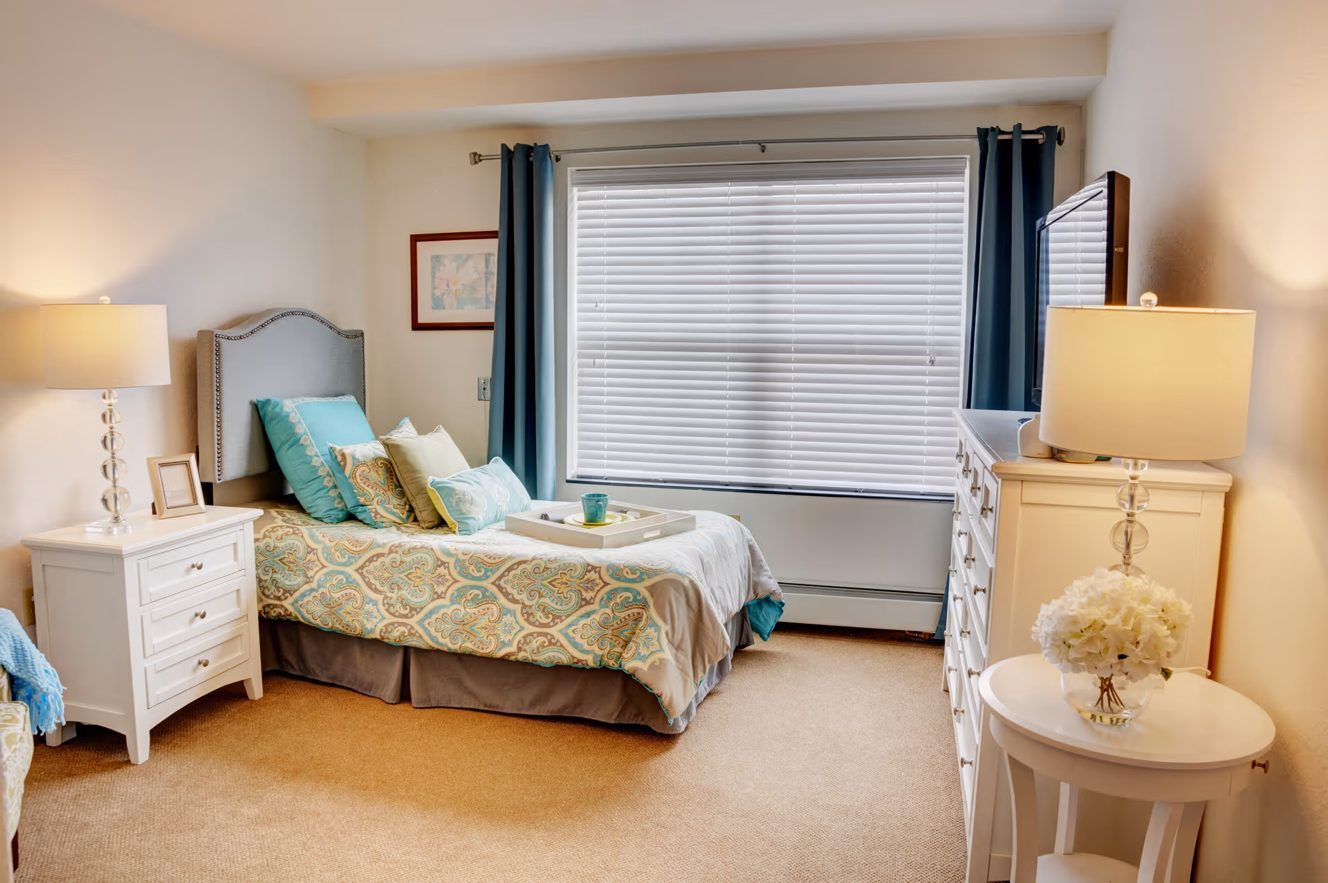 Sunlit bedroom with a single bed dressed in patterned bedding, bedside tables and lamps, a dresser, and a large window with blinds.
