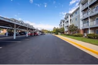 View of a paved parking area with covered parking spaces on the left and a multi-story residential building with balconies on the right under a partly cloudy blue sky.