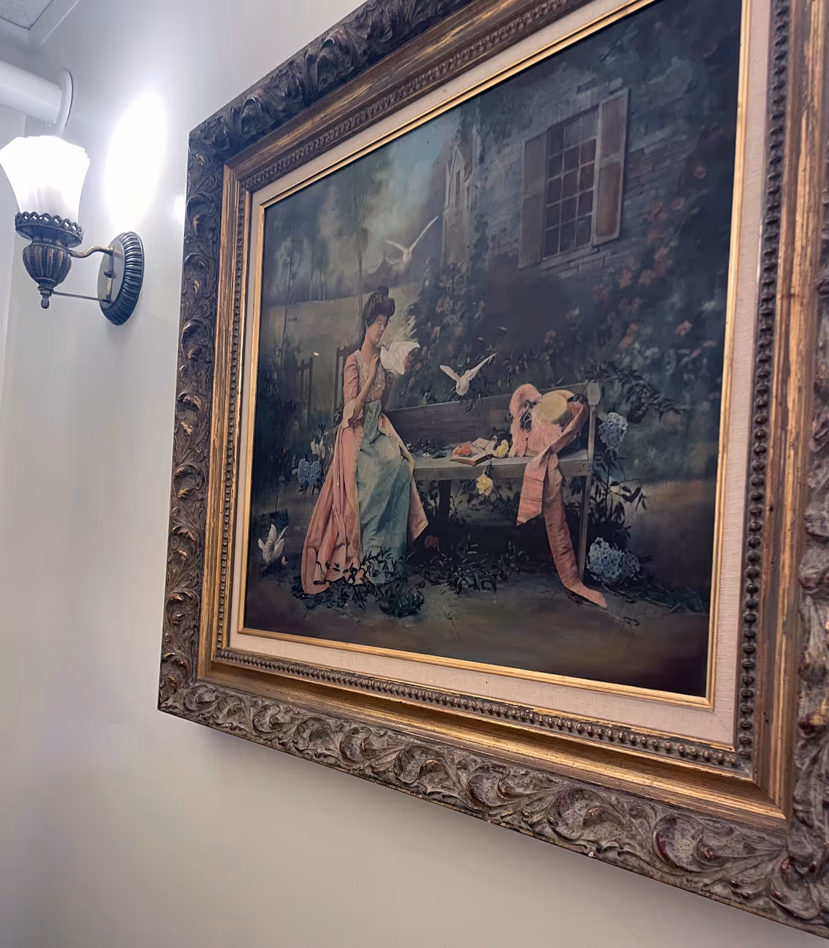 A framed painting on a wall depicting a woman in a long dress sitting on a bench in a garden setting with birds flying around her. The painting has an ornate frame and is illuminated by a wall-mounted light fixture with a frosted glass shade.