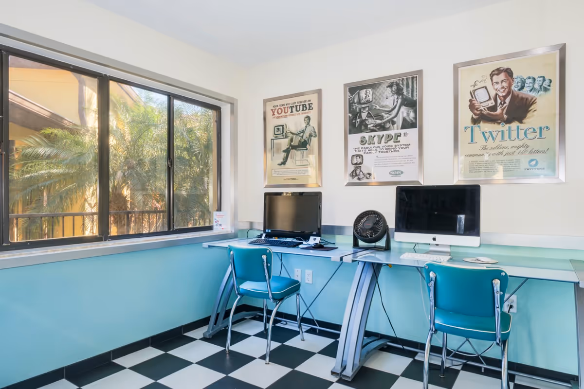 A small computer room with two desks, each equipped with a computer and a teal chair. The room has a large window showing palm trees outside. The walls are painted light blue and white, and there are three framed vintage-style posters on the wall advertising YouTube, Skype, and Twitter. The floor has a black and white checkered pattern.