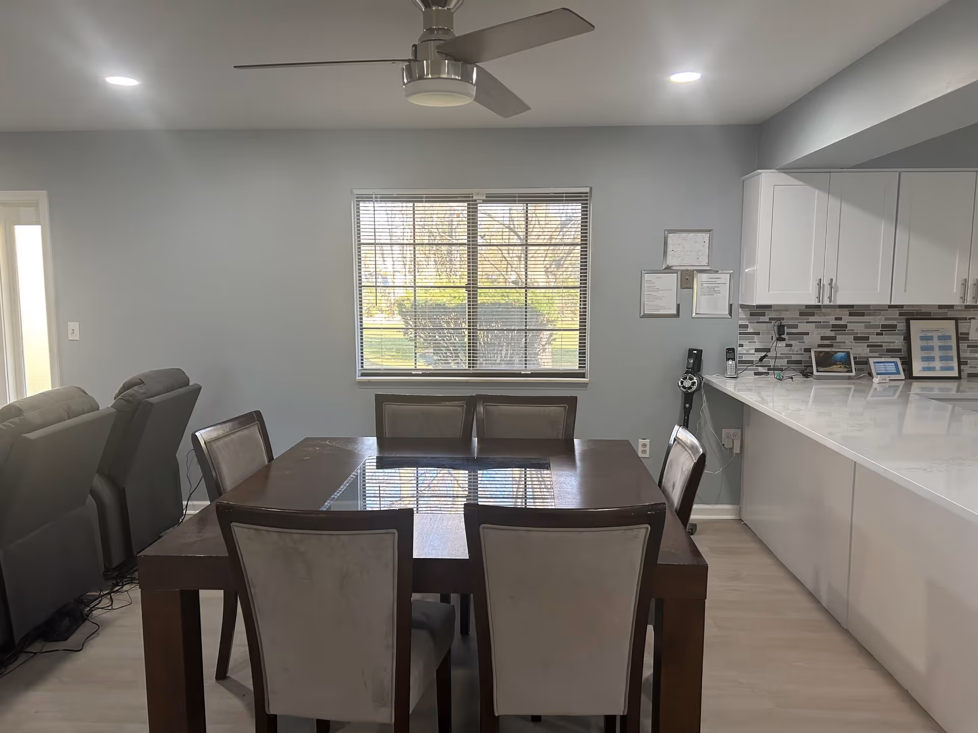 A modern dining area with a dark wooden table and six upholstered chairs. Behind the table is a large window with blinds partially open, showing greenery outside. To the right is a kitchen counter with white cabinets and a tiled backsplash. On the counter are several electronic devices and framed documents. On the left side, there are two gray recliner chairs. The ceiling has recessed lighting and a ceiling fan with three blades.