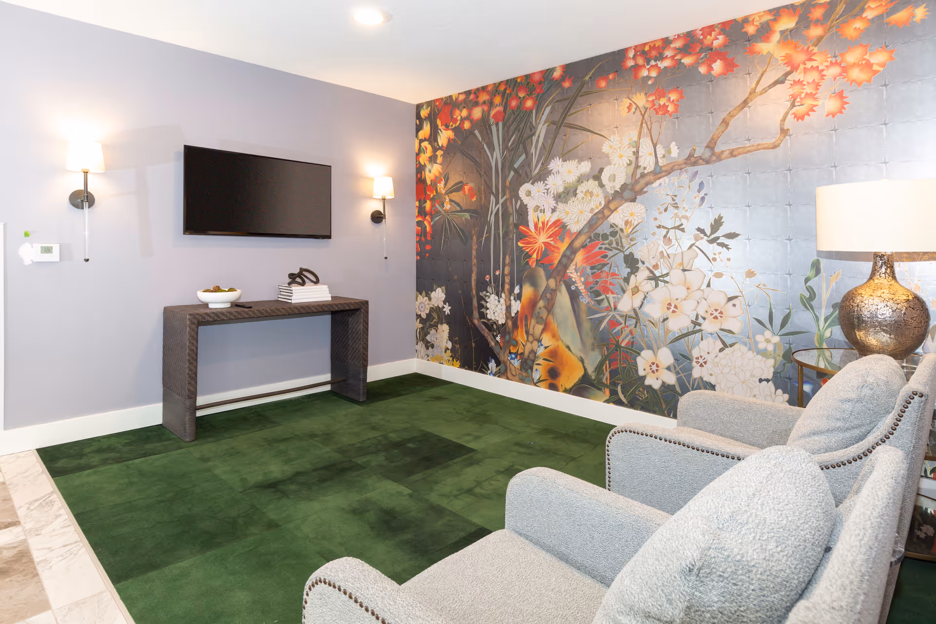 Seating area with two upholstered chairs facing a wall-mounted TV, a console table, lamp, and a large floral mural on the wall.