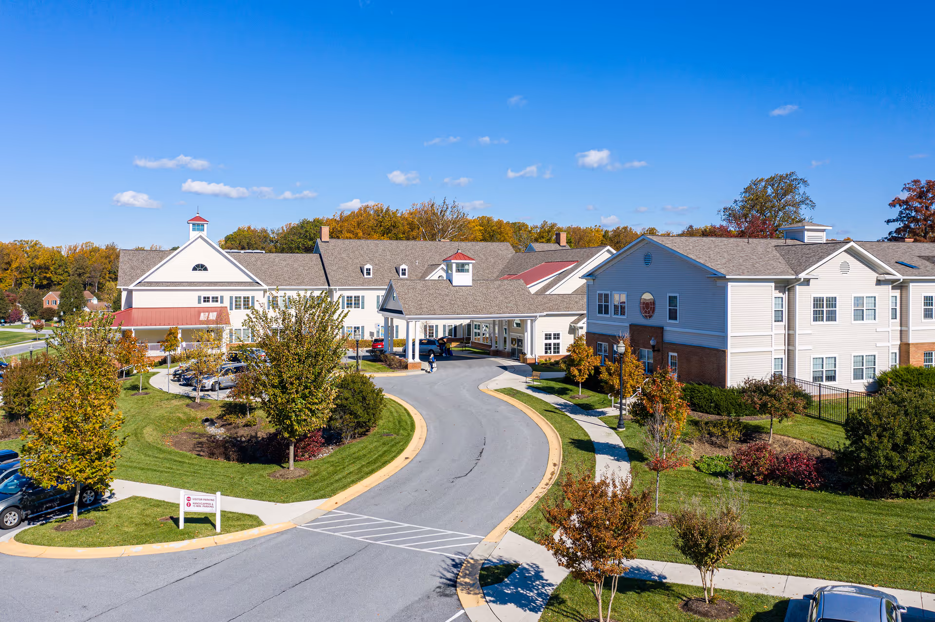 Aerial view of a large senior care facility with a covered entrance, circular driveway, landscaped lawns and trees under a clear blue sky.