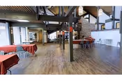 Spacious interior common area with wooden flooring, exposed beams, and large windows. Several tables covered with red and white checkered tablecloths are arranged throughout the room, with chairs around them. A stone fireplace is visible in the background, along with modern lighting fixtures and a high ceiling.