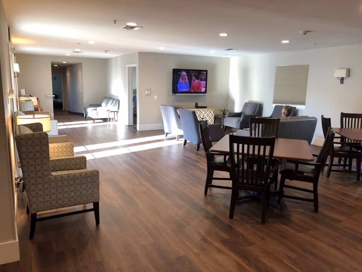 A spacious senior living common area with wooden flooring, several armchairs and tables with chairs. A television is mounted on the far wall showing a program with two people. There is a person sitting on a couch watching the TV. The room is well-lit with ceiling lights and natural light coming from windows.