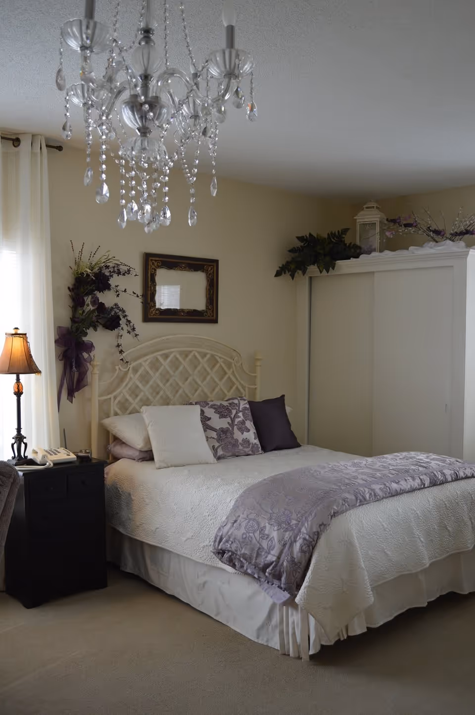 A cozy bedroom with a white bed featuring a quilted bedspread and purple floral pillows. A black nightstand with a lamp and a telephone is beside the bed. Above the bed is a decorative mirror and floral wall decor. A white wardrobe with sliding doors is in the corner, topped with plants and a lantern. A crystal chandelier hangs from the ceiling.