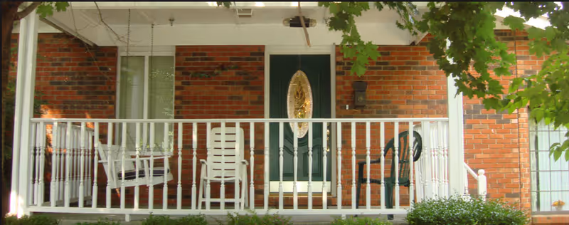 Front porch of a brick building with a green door featuring an oval glass panel. The porch has white railings and several chairs, including a white rocking chair, a white chair, and a dark green chair. There are green leaves from a tree partially visible in the foreground.