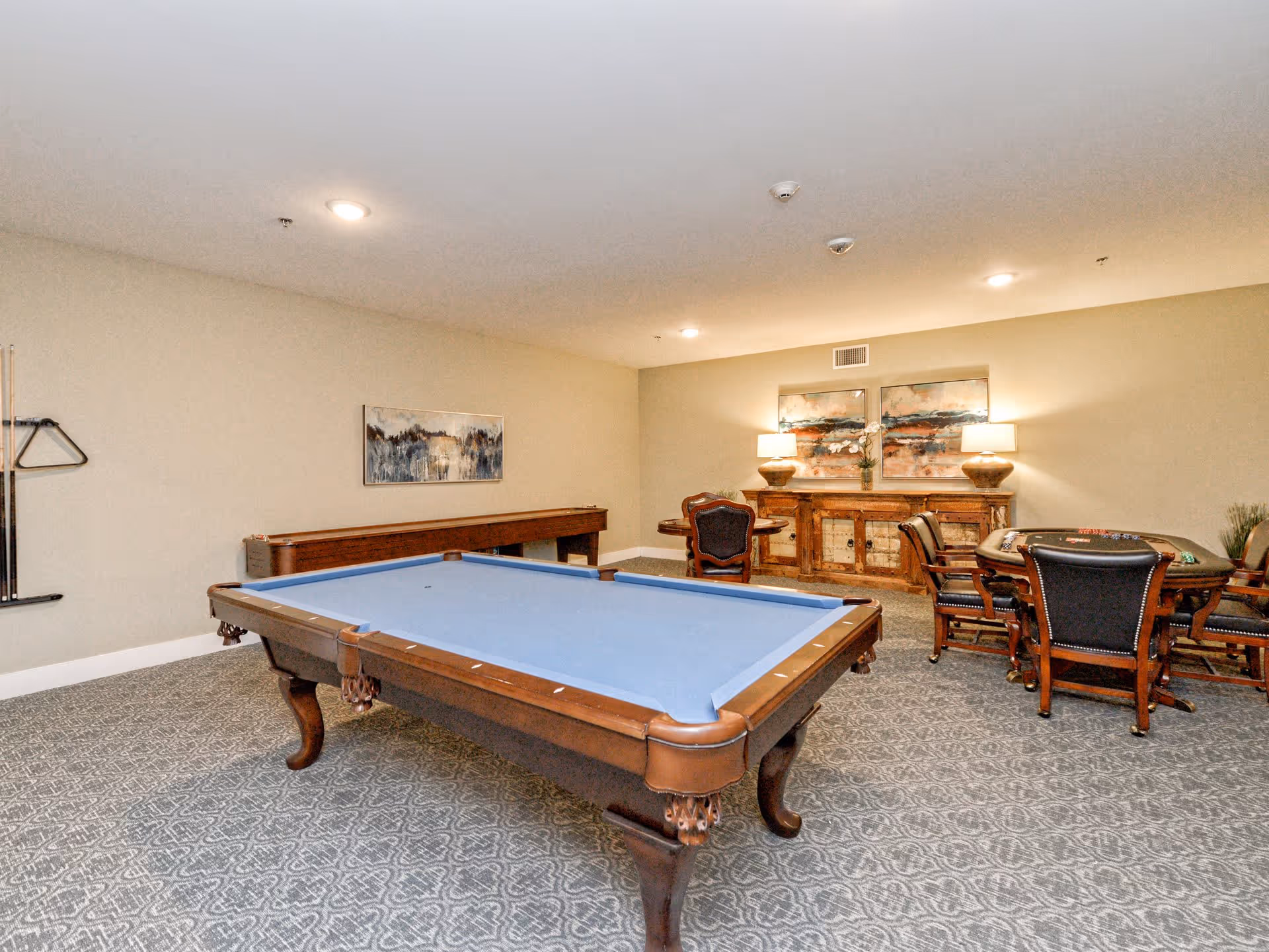 A recreational room with a blue felt pool table in the center, a shuffleboard table against the wall, and a poker table with chairs. The room has patterned carpet, beige walls, two table lamps on a wooden sideboard, and two abstract paintings above the sideboard.