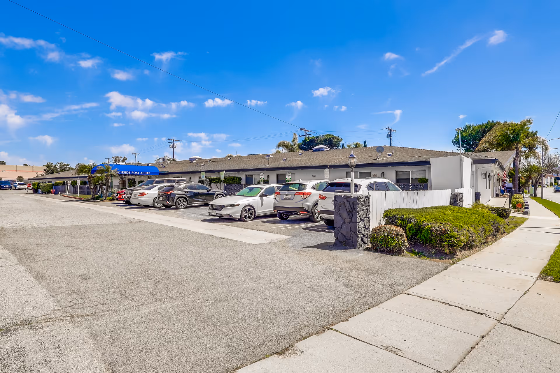 Exterior view of Beachside Post Acute facility showing a single-story building with a parking lot filled with cars in front. The sky is clear with a few clouds, and there are some palm trees and shrubs around the building.