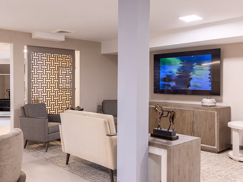 A modern living room area with beige and gray armchairs and sofas arranged around a wooden coffee table. A flat-screen TV is mounted on the wall above a wooden cabinet, and a decorative horse sculpture is placed on a side table. The room features neutral tones and a patterned room divider in the background.