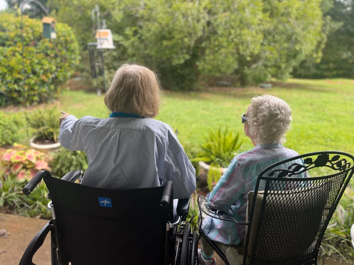 Two elderly women sitting outside in a garden area, one in a wheelchair and the other in a metal chair, facing away from the camera and looking at greenery and plants.