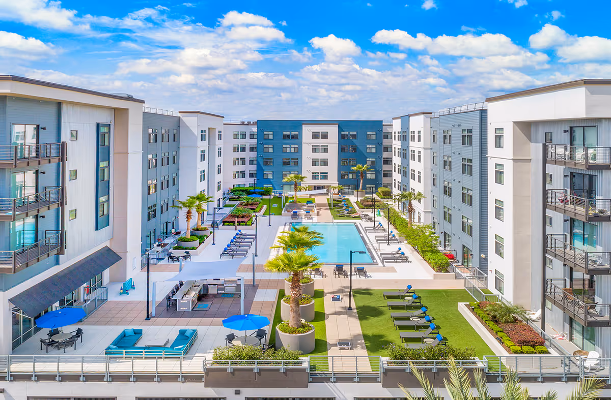 Outdoor courtyard area of a modern residential facility with a swimming pool in the center, surrounded by lounge chairs, palm trees, seating areas with blue umbrellas, and multi-story buildings on all sides under a partly cloudy blue sky.