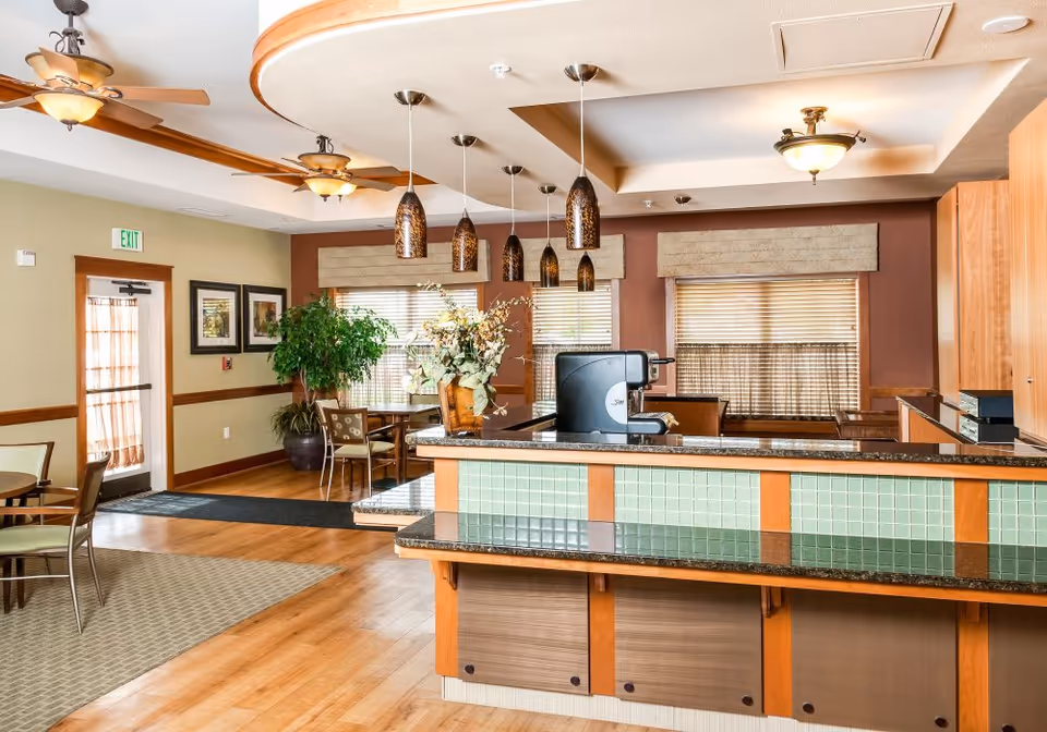 A bright and spacious common area in a senior living facility featuring a counter with green tiles and a coffee machine, pendant lights hanging from the ceiling, wooden flooring, several tables and chairs, large windows with blinds, and decorative plants.