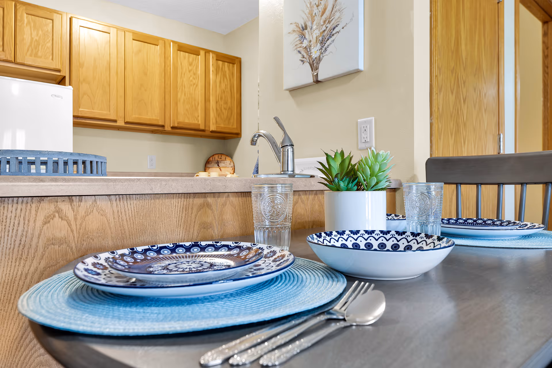 A dining table set for two with blue patterned plates, clear drinking glasses, silverware, and a small white pot with green succulent plants. In the background, there is a kitchen counter with wooden cabinets, a faucet, and a wall painting featuring dried plants.