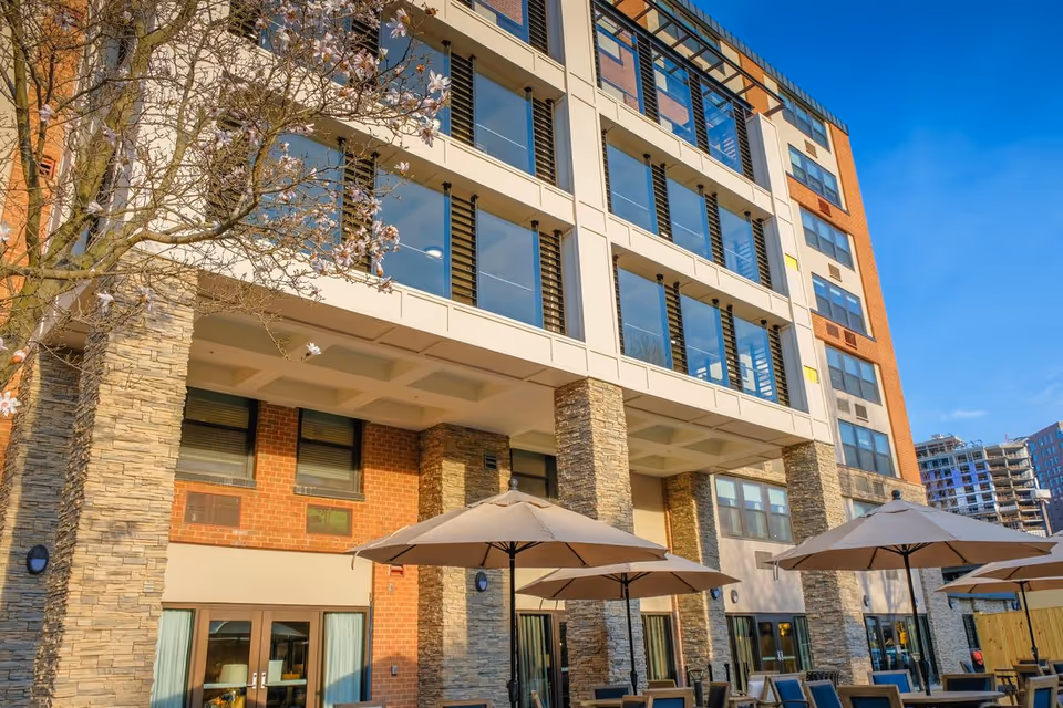 Front exterior of a multi-story residential building with stone pillars, large windows, outdoor patio tables with umbrellas, and a flowering tree.