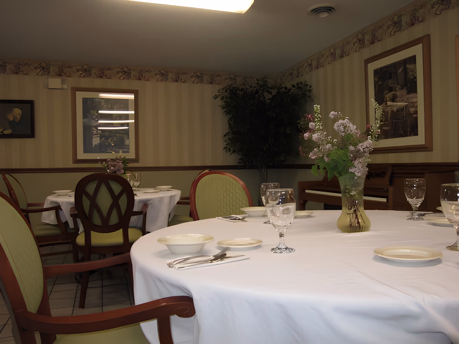 A dining room with round tables covered in white tablecloths, set with plates, bowls, silverware, and glasses. There is a vase with flowers on the table in the foreground. The room has patterned wallpaper, framed artwork on the walls, and a piano in the background.