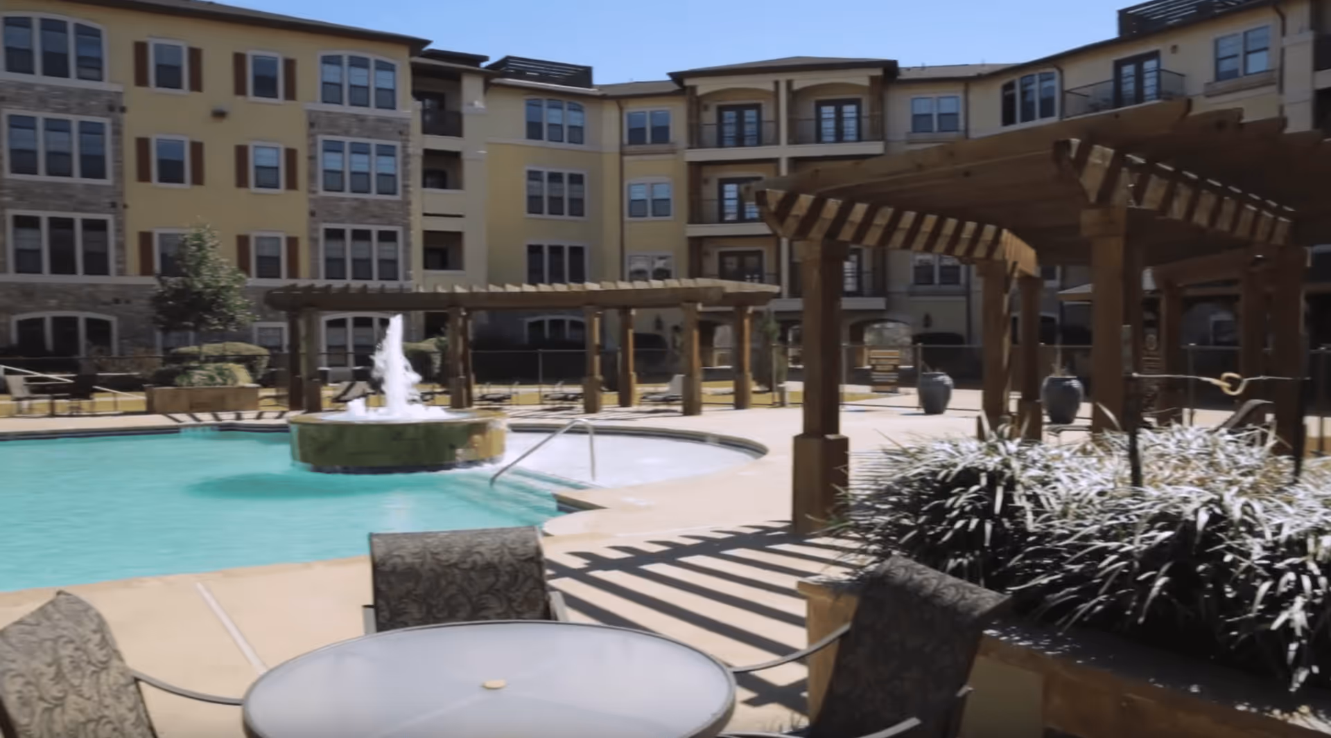 Outdoor courtyard area of a senior living facility featuring a swimming pool with a central fountain, wooden pergolas, patio tables with chairs, and landscaped plants. The multi-story building with numerous windows surrounds the courtyard.