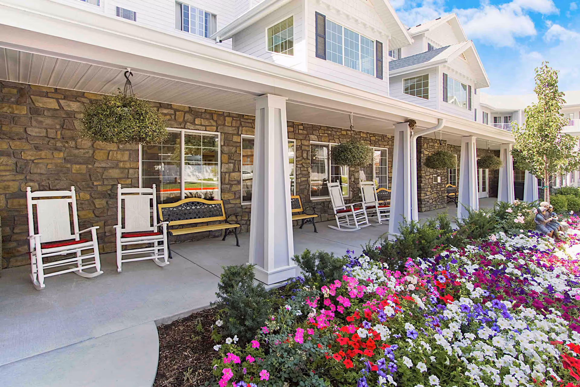 Covered front porch of a senior living residence with rocking chairs, hanging baskets, and a colorful flowerbed.