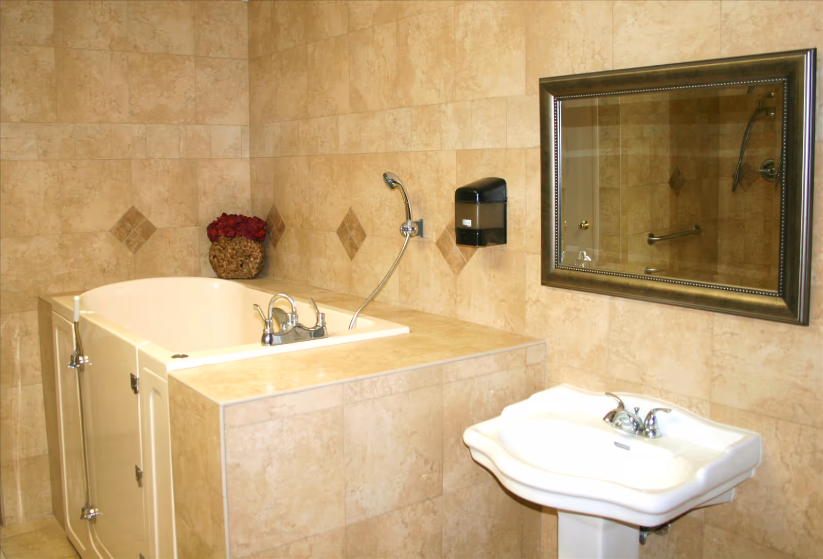 A bathroom with beige tiled walls featuring diamond-shaped accents, a walk-in bathtub with a handheld showerhead, a white pedestal sink with chrome faucets, a large framed mirror above the sink, and a decorative basket with red flowers placed on the bathtub ledge.