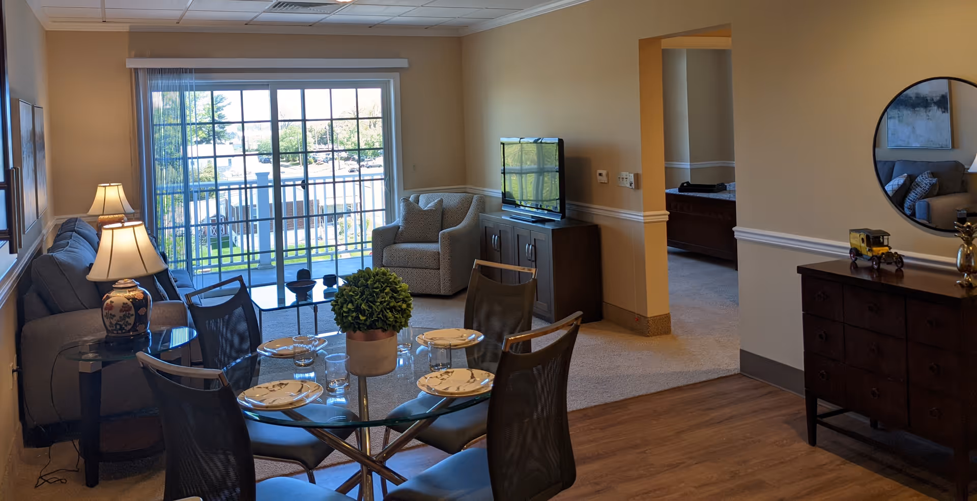 A cozy living and dining area in a senior living facility with a glass dining table set for four, a sofa, an armchair, a TV on a stand, a lamp on a side table, and a large sliding glass door leading to a balcony with a view of greenery outside. There is a wooden dresser with a round mirror above it and decorative items on top.
