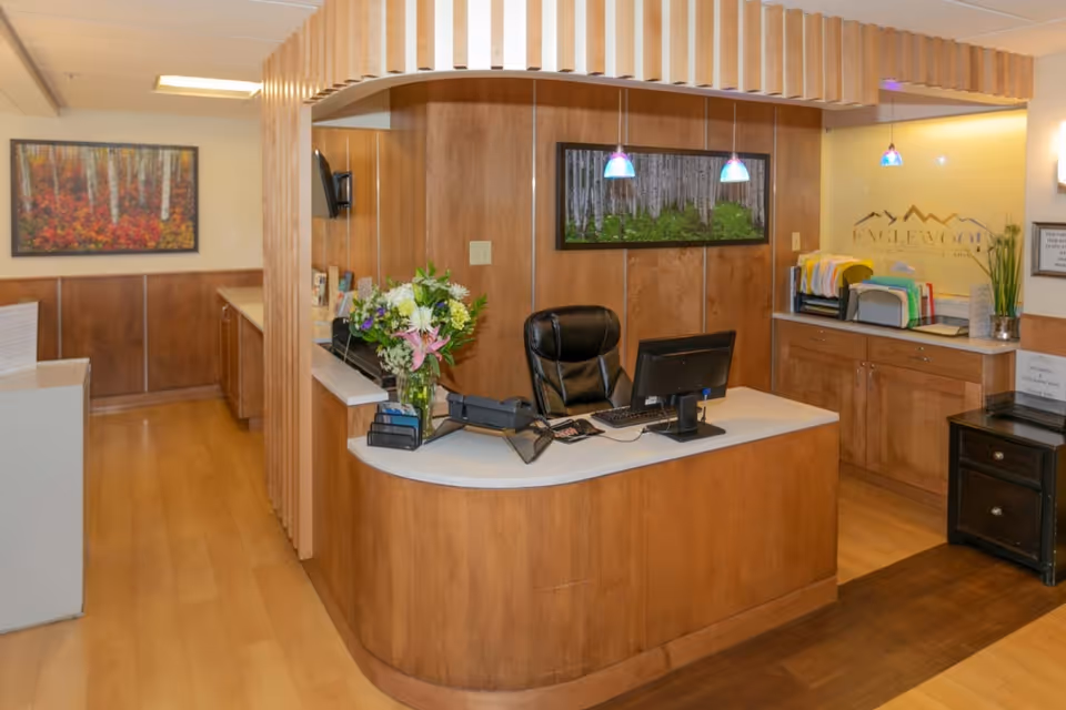 Reception area with a wooden curved desk, a black office chair, a computer monitor, and a vase of flowers. The background features wood-paneled walls, two framed pictures of forest scenes, hanging pendant lights, and filing trays on a countertop. The facility name 'Englewood Post Acute and Rehabilitation' is visible on a glass panel behind the desk.