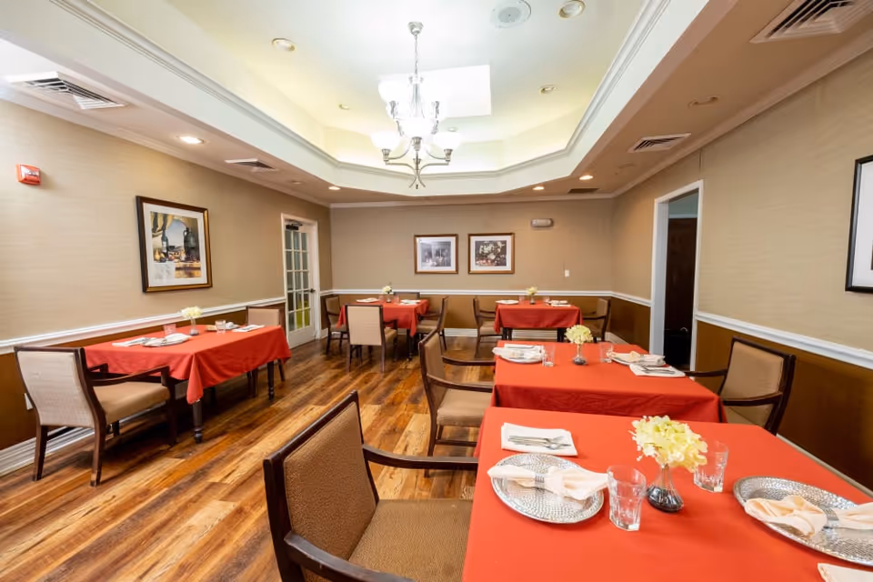 Bright dining room with multiple tables covered in red tablecloths, place settings, chairs, and a central chandelier.