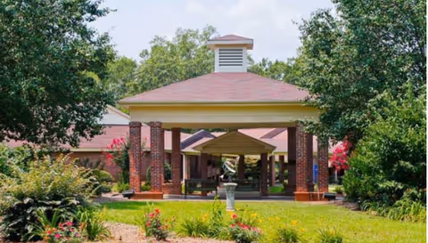 Exterior view of a senior living facility entrance with a covered driveway supported by brick columns, surrounded by green trees and landscaped garden beds with flowers.