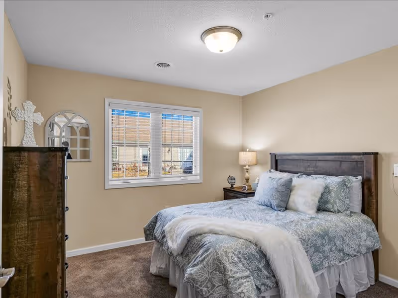 A cozy bedroom with beige walls and carpeted floor featuring a bed with a wooden headboard, light blue and white patterned bedding, and multiple pillows. There is a wooden nightstand with a lamp and a small clock beside the bed. A window with white blinds lets in natural light, and a tall wooden dresser with decorative items is positioned against the left wall.